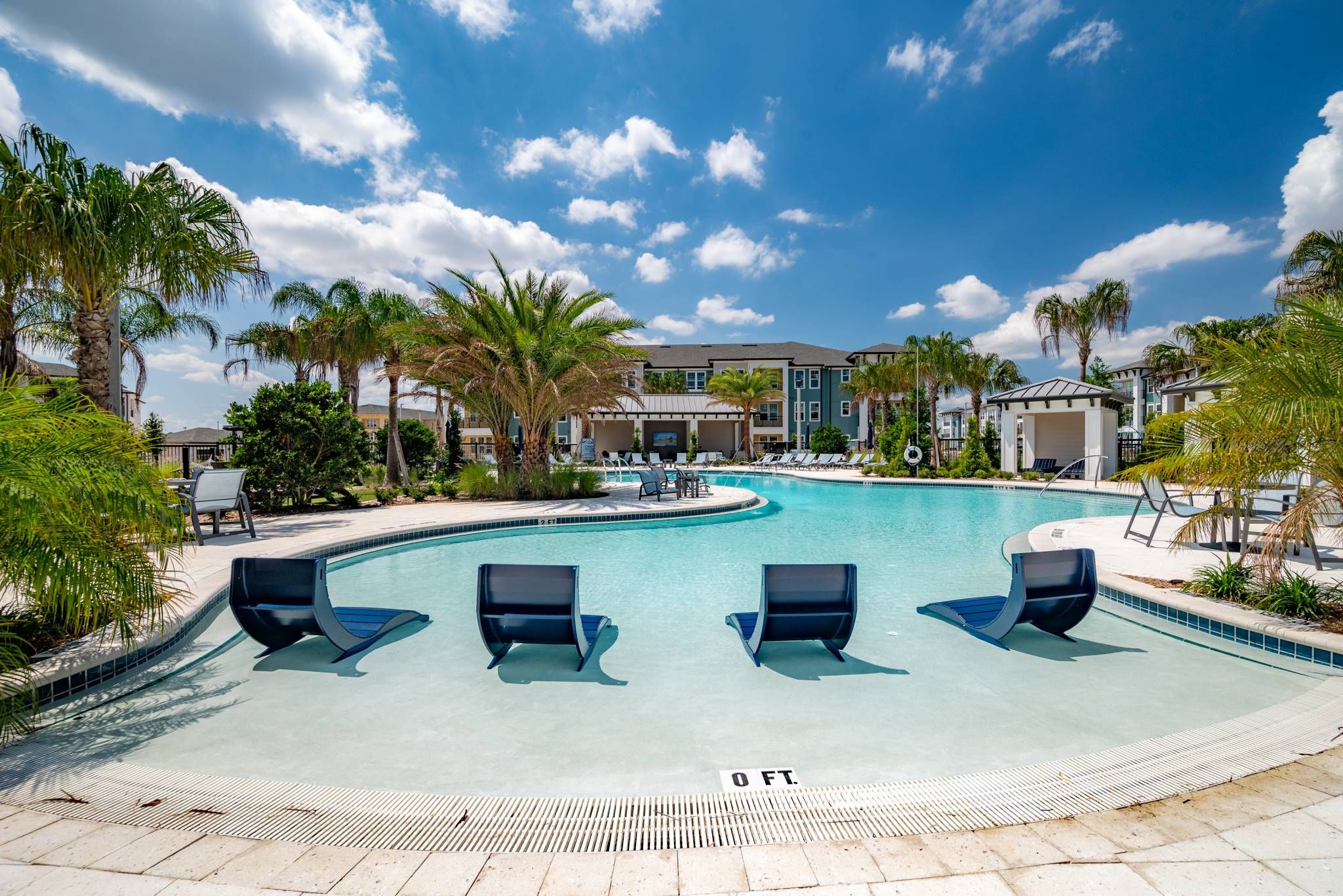 An inviting zero-entry swimming pool with lounge chairs partially submerged in the water, surrounded by palm trees and apartment buildings at Alta at Horizon West.