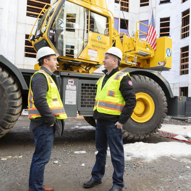 Two construction workers stand in front of a large bulldozer at a construction site.