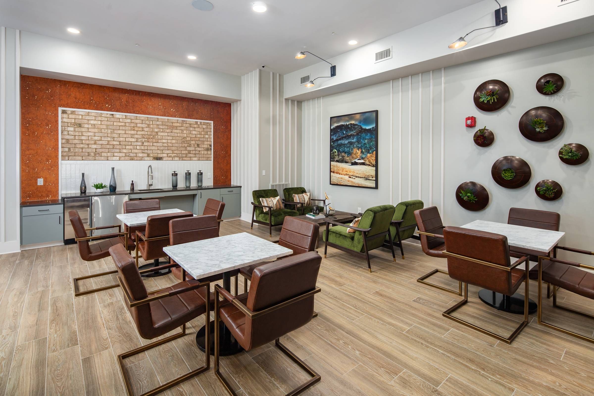 Alta Sugarloaf's dining space with leather chairs, marble-top tables, and a kitchenette against a brick backsplash, complemented by vertical greenery and modern artwork.