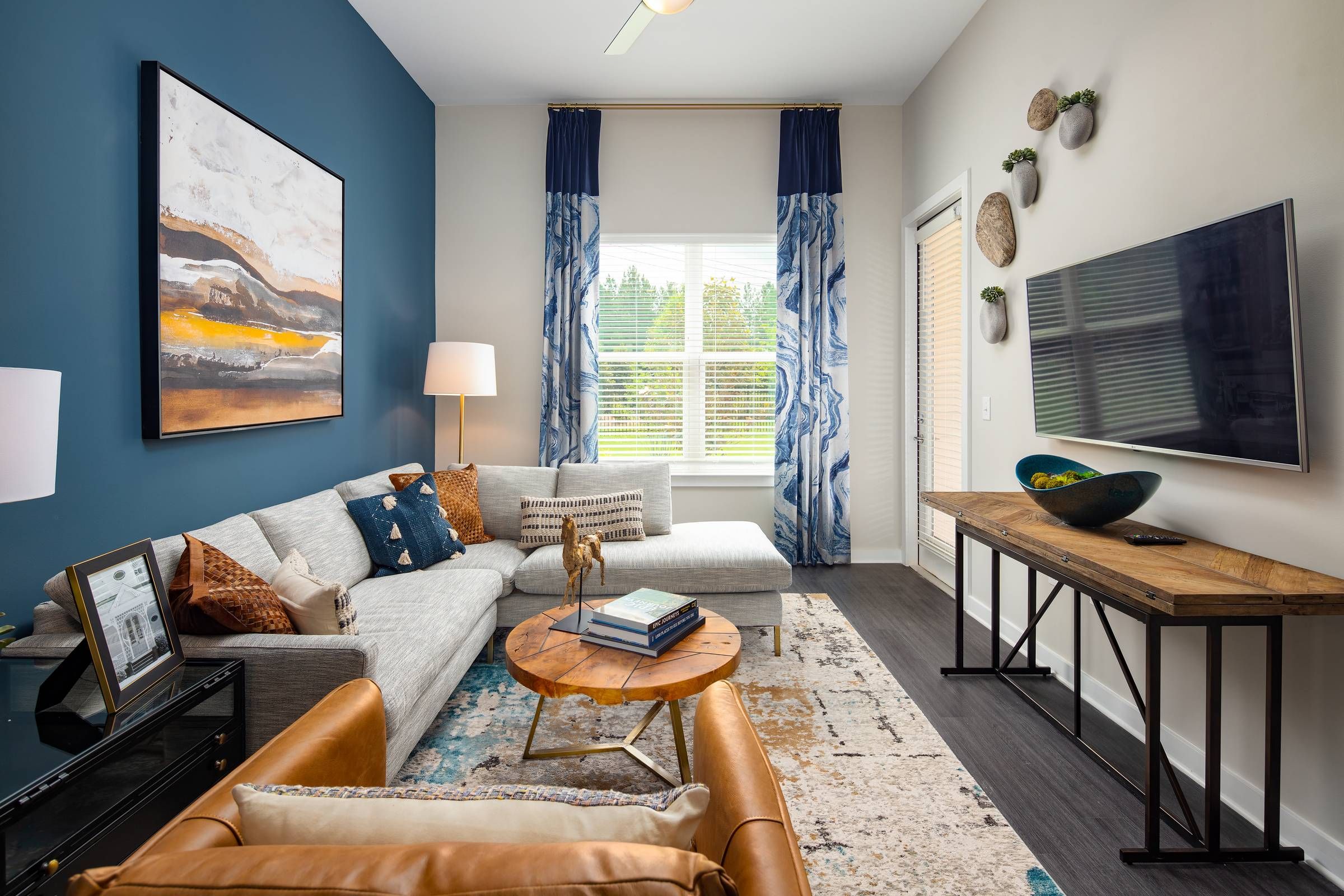 A stylish living room at Alta Sugarloaf with a gray sectional sofa, blue accent wall, eclectic decor, and large windows draped in patterned curtains.