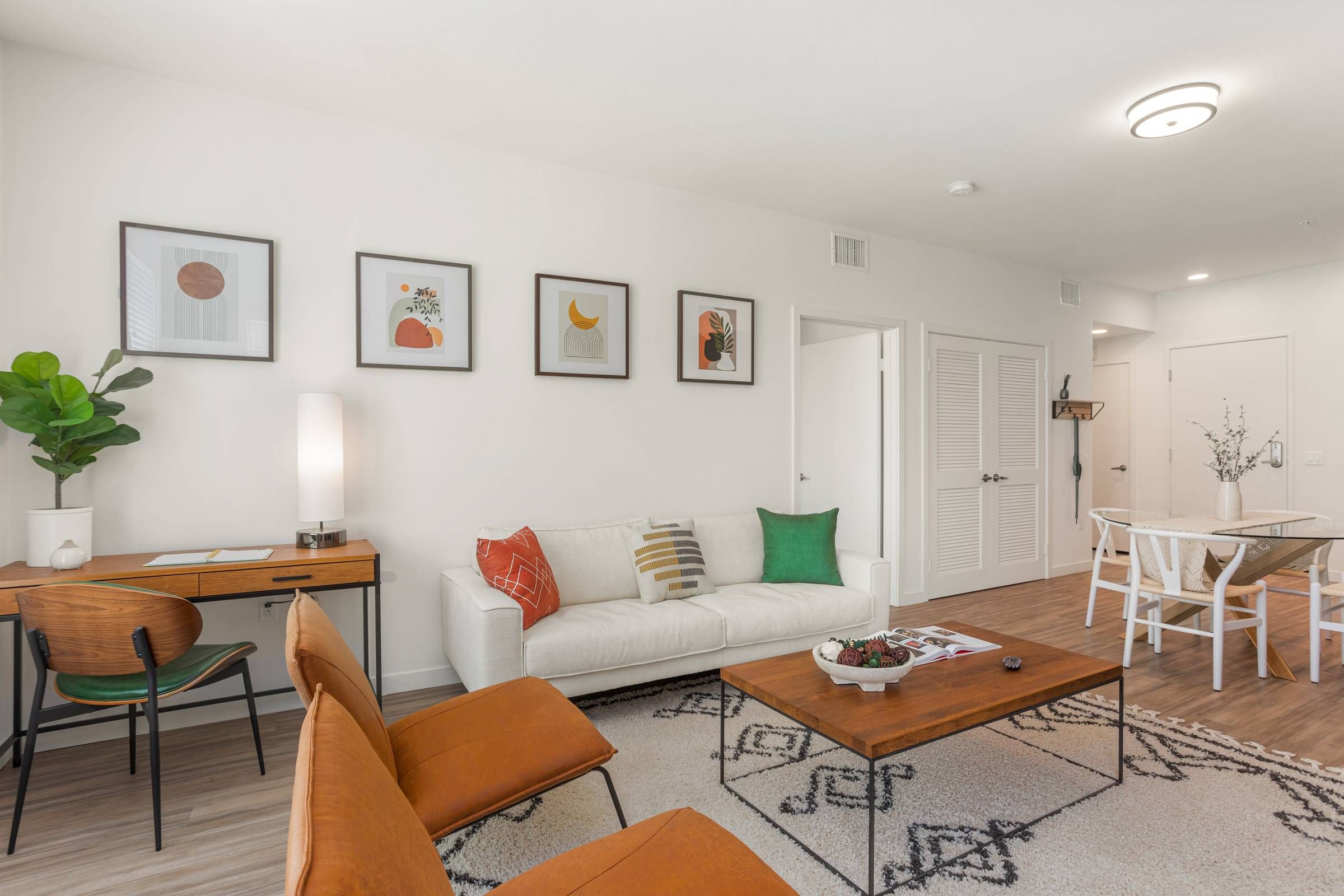 Alta Upland presents a bright living room adorned with colorful wall art above a minimalistic desk, a green potted plant, and mid-century modern furnishings.