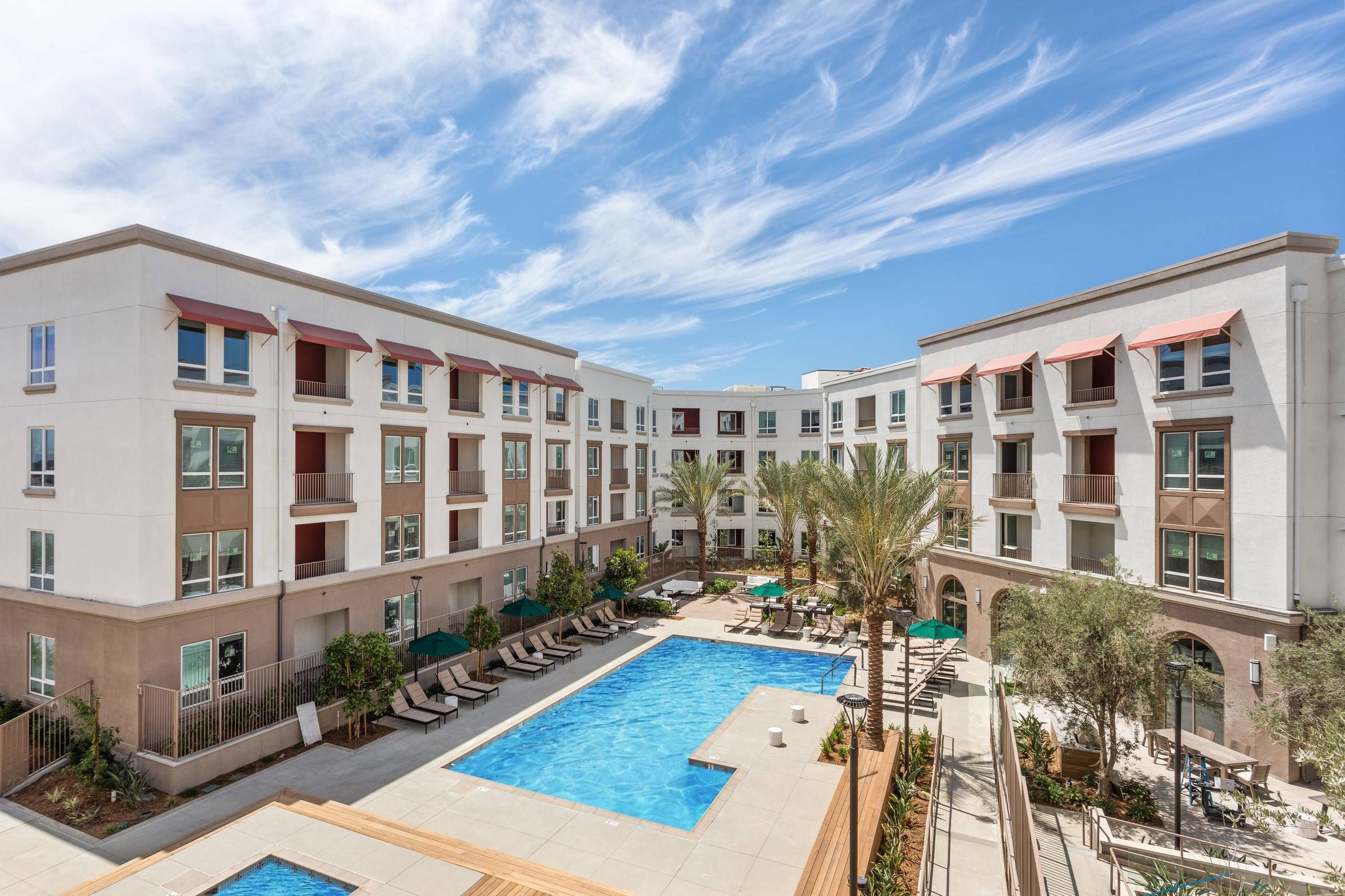 Alta Upland's luxurious outdoor pool is surrounded by sun loungers and palm trees, nestled within a modern apartment complex.