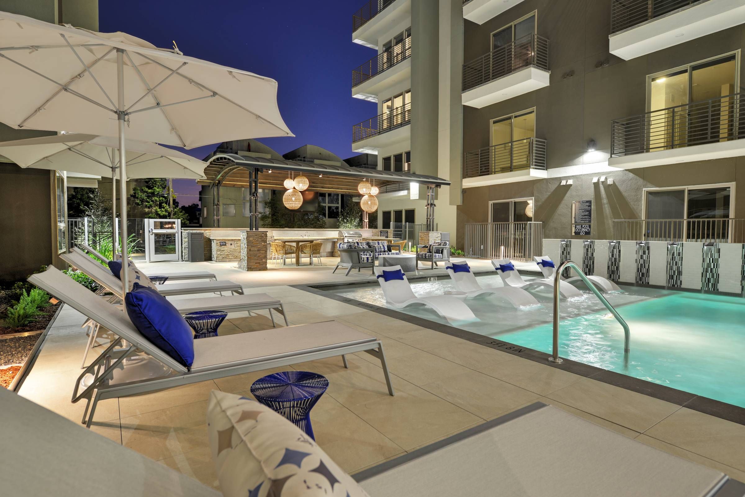 A tranquil evening pool scene at Alta West Gray, with white loungers under umbrellas and a warmly lit outdoor kitchen and dining area in the background.