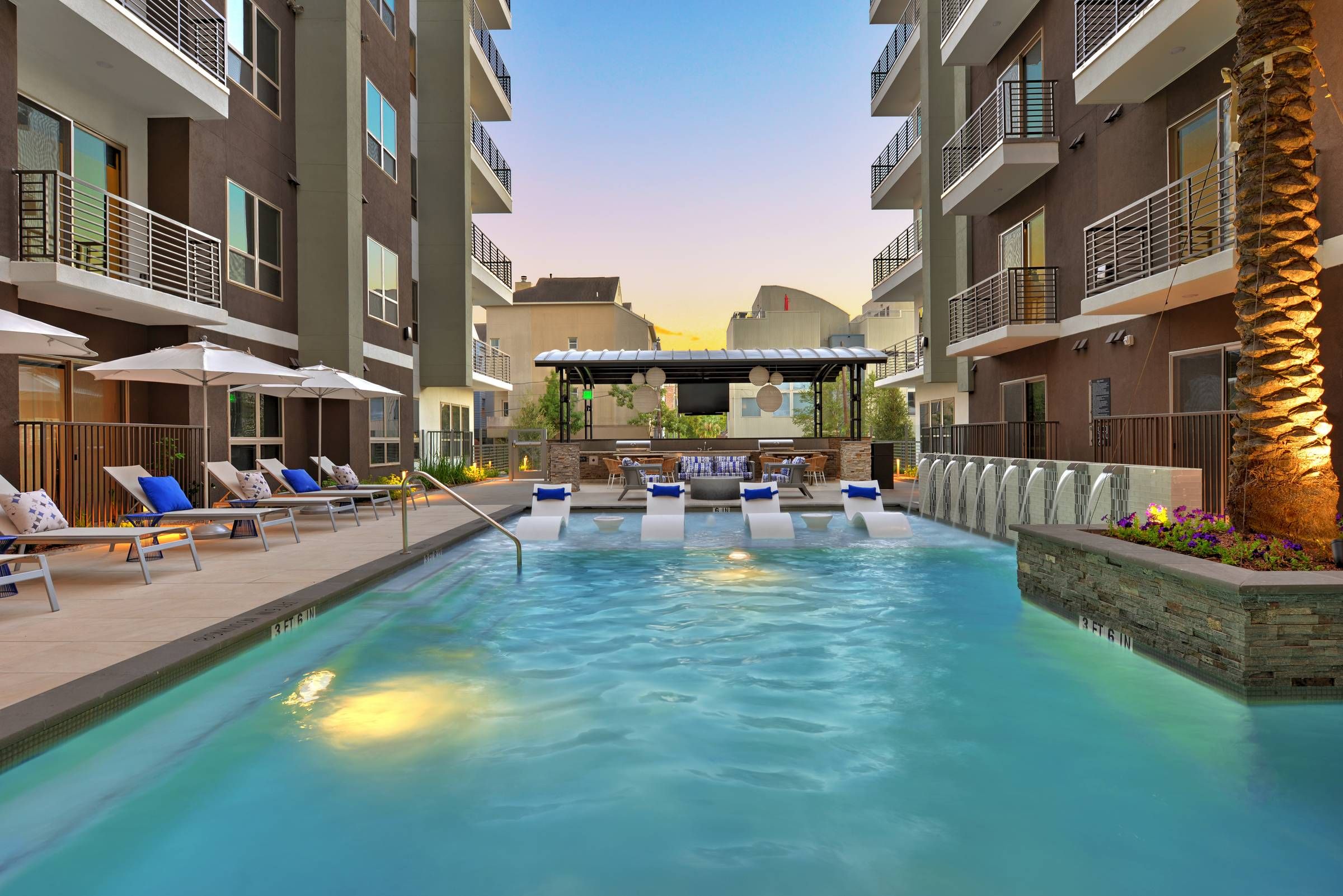 Alta West Gray's pool area with loungers, blue umbrellas, and an outdoor kitchen, surrounded by multi-story residential buildings at sunset.