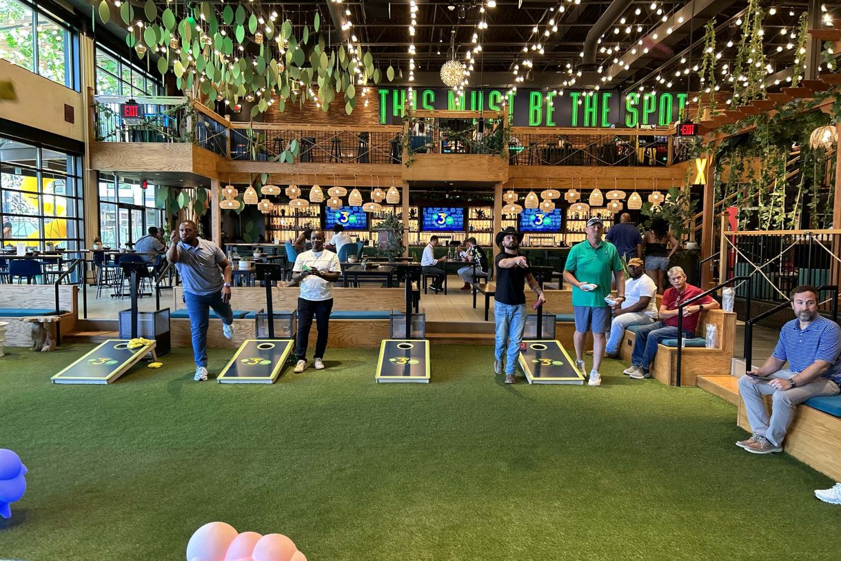 People playing cornhole on astroturf inside of decorated building used for cornhole tournament