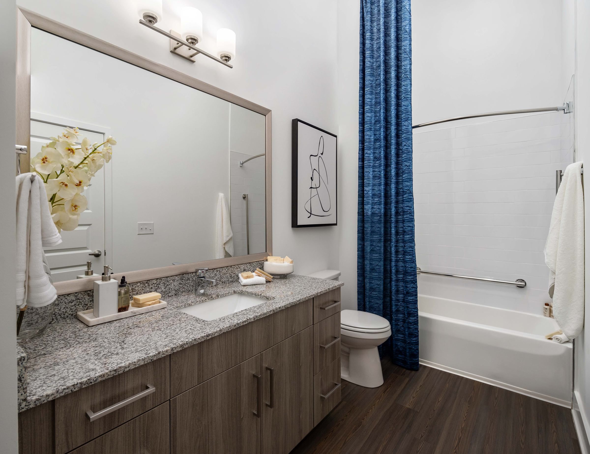 The bathroom at Alta Ashley Park, featuring a granite countertop, large mirror, and a blue shower curtain that adds a pop of color to the neutral-toned space.
