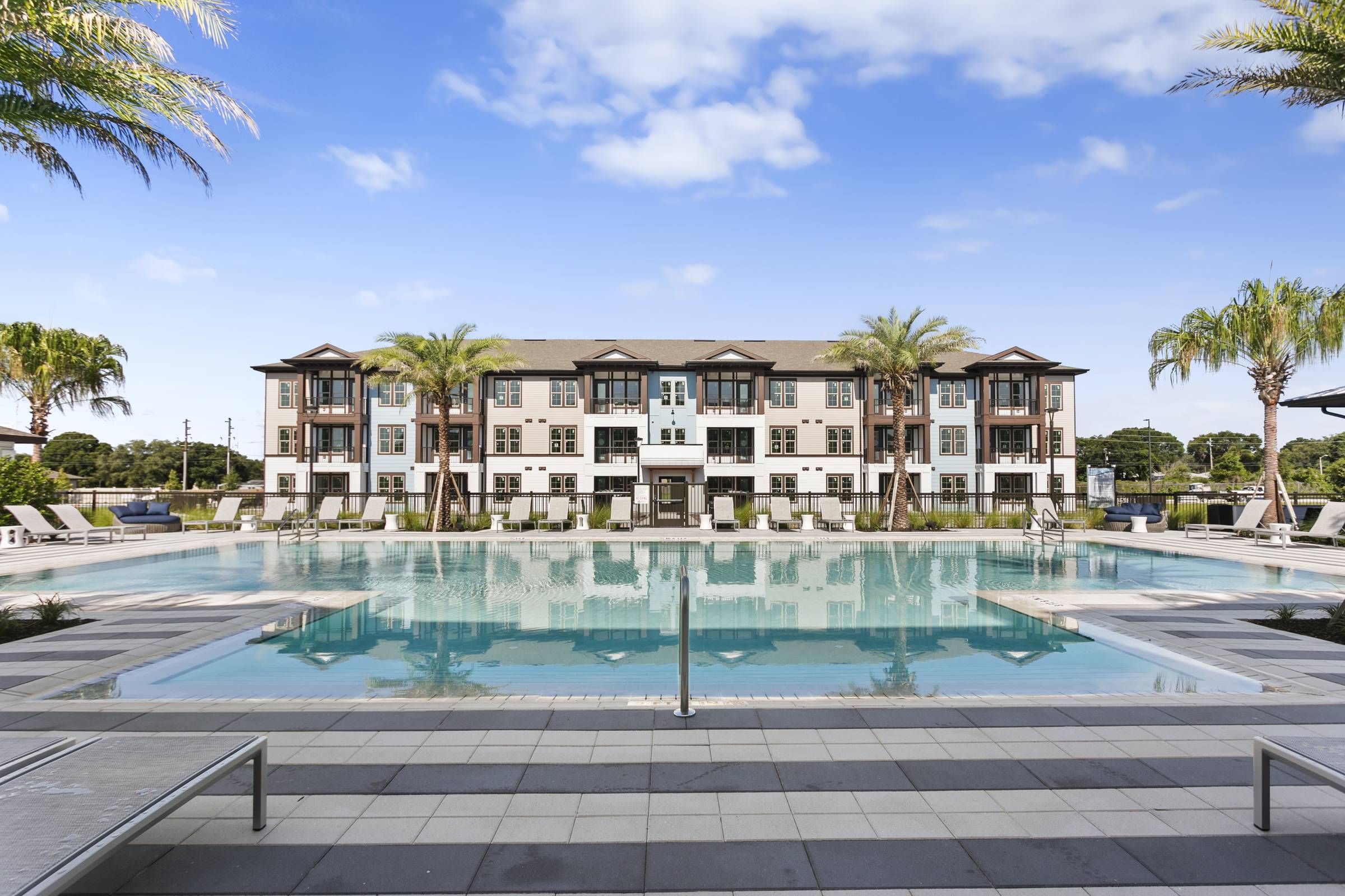 Alta Clearwater's serene poolside view showcases lounging chairs facing a large, reflective swimming pool, flanked by a stately three-story building with palm trees.