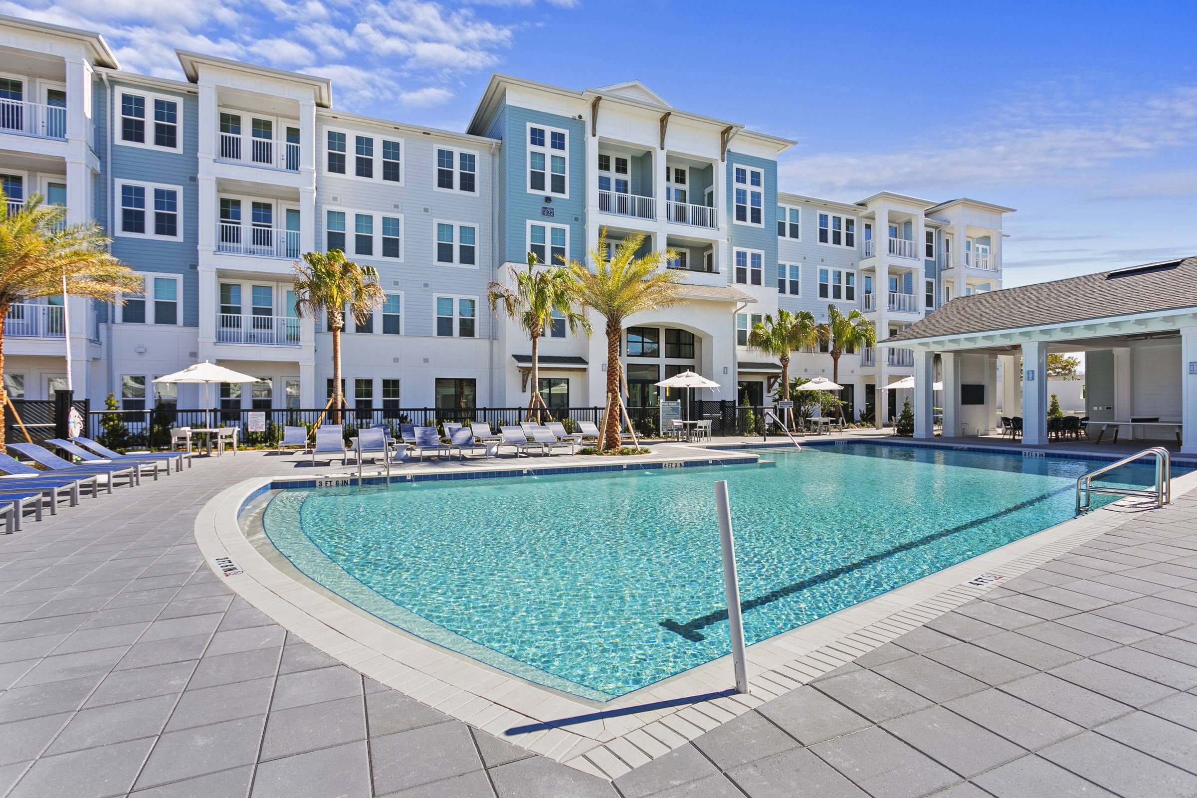 A sparkling blue swimming pool at Alta Belleair flanked by a white multi-story building and a row of sun loungers, set under a clear blue sky.
