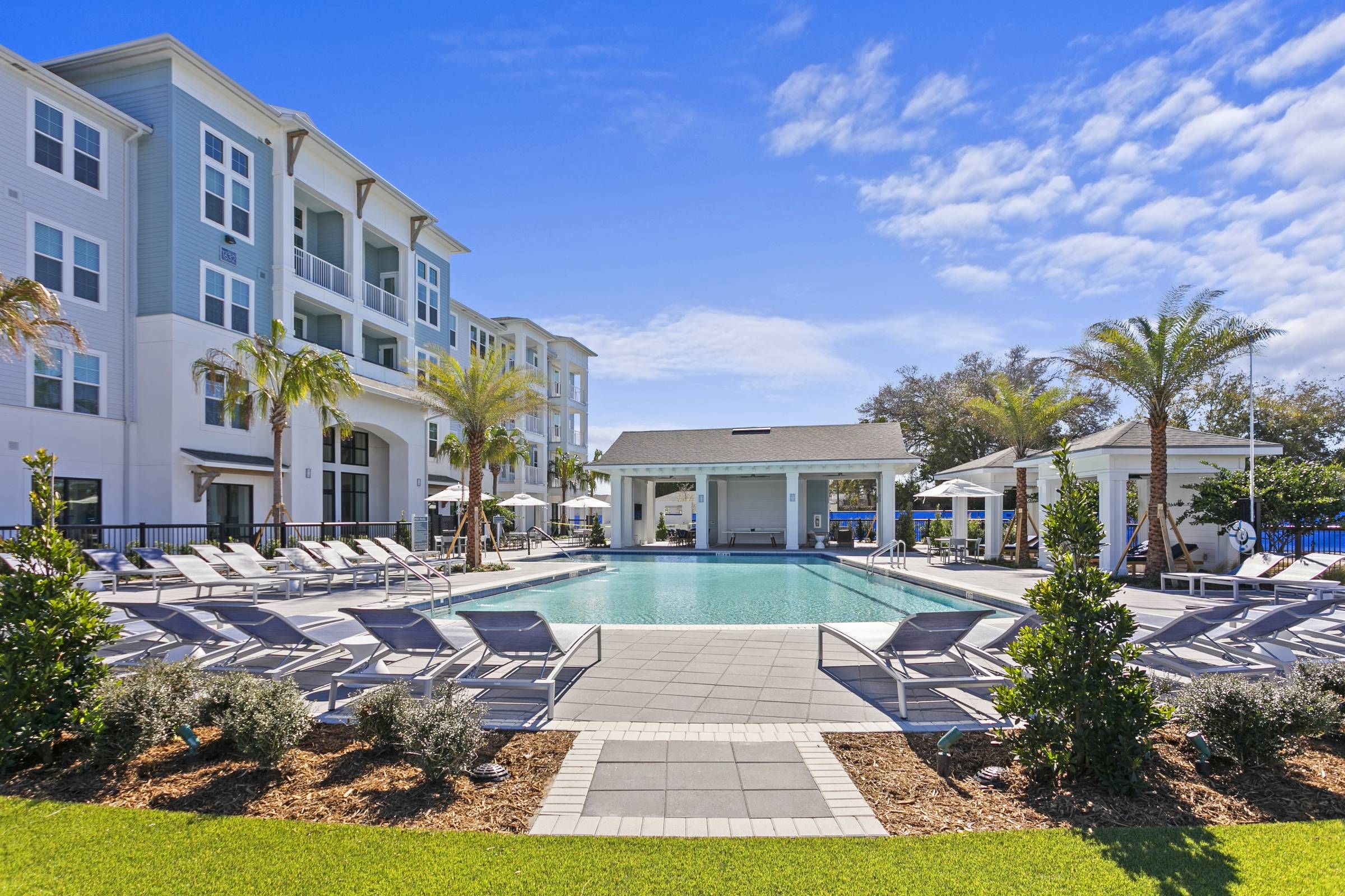 The inviting pool area at Alta Belleair is framed by stylish white loungers, green landscaping, and a contemporary outdoor pavilion under a sunny sky.
