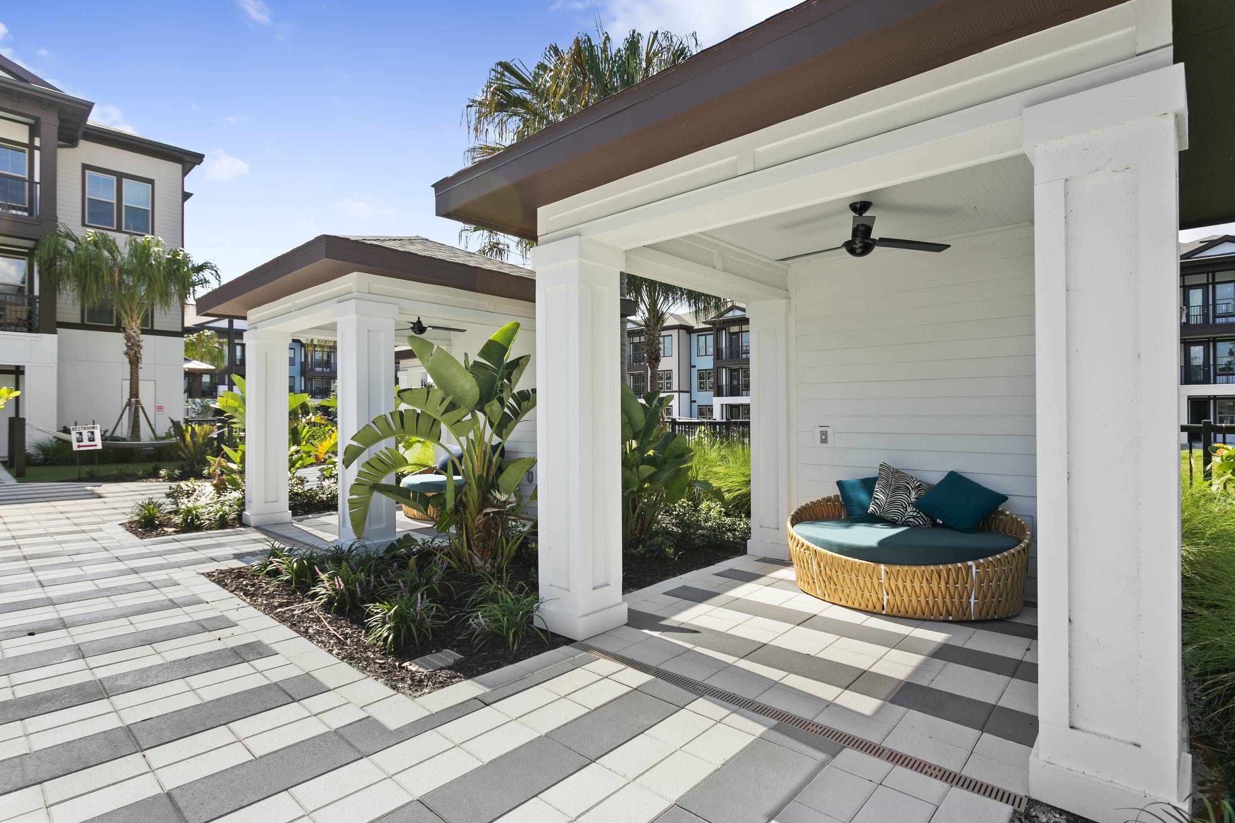 An outdoor lounge area at Alta Clearwater with a circular rattan sofa under a pergola, amidst lush tropical landscaping and checkerboard walkways.