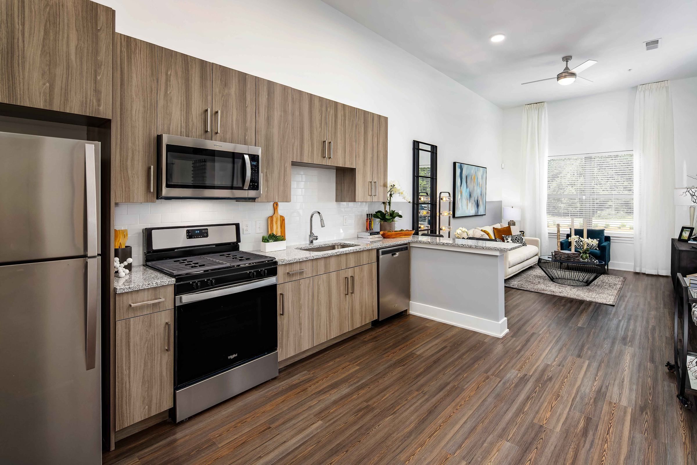 A chic kitchen in Alta Ashley Park, with sleek gray cabinets, stainless steel appliances, and a cozy living area illuminated by natural light from large windows.