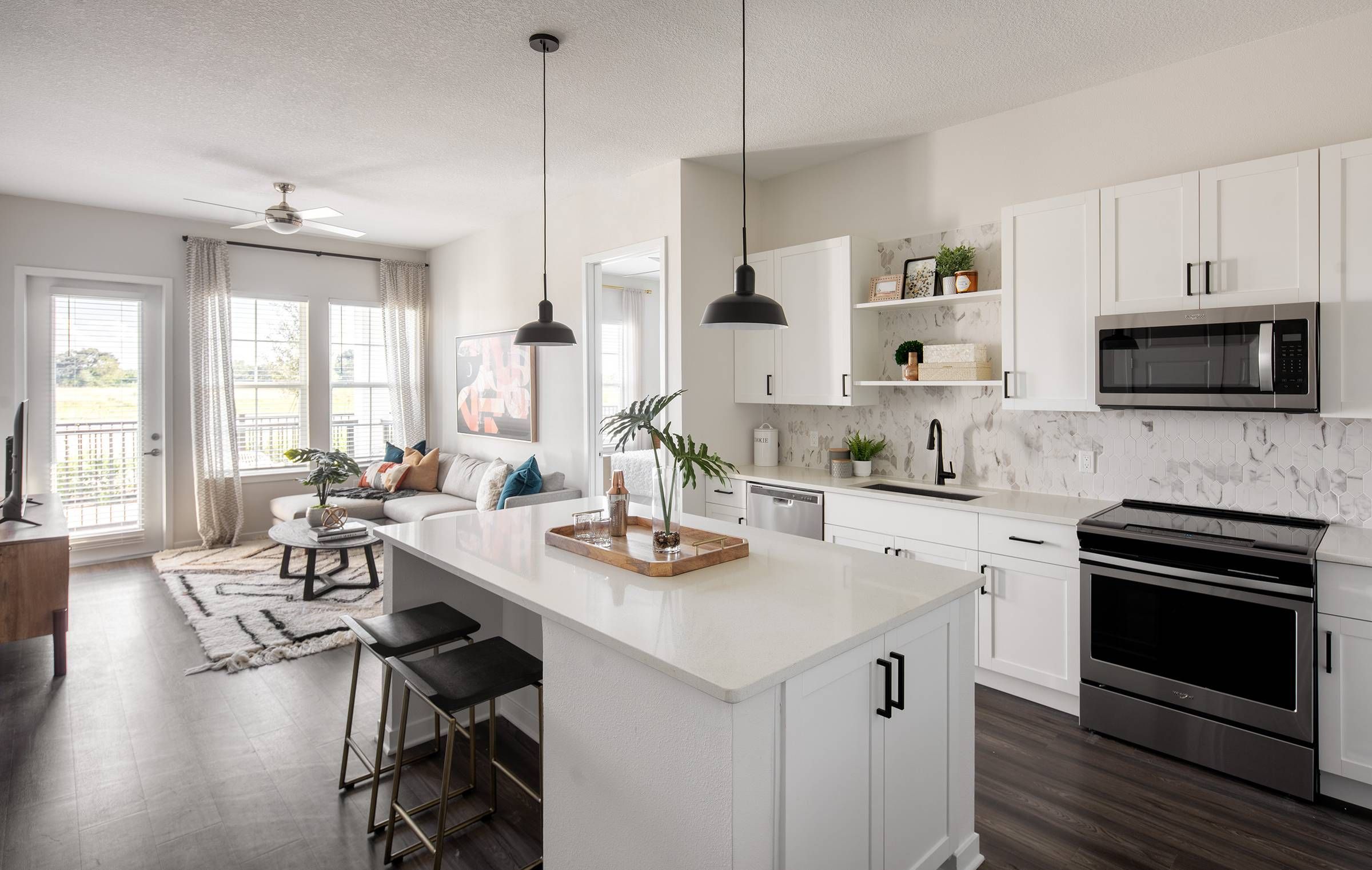 Alta Winter Garden apartment's open-concept two-bedroom model showcasing a kitchen with white cabinetry and marble backsplash, leading into a cozy living area with a sectional sofa and balcony access.