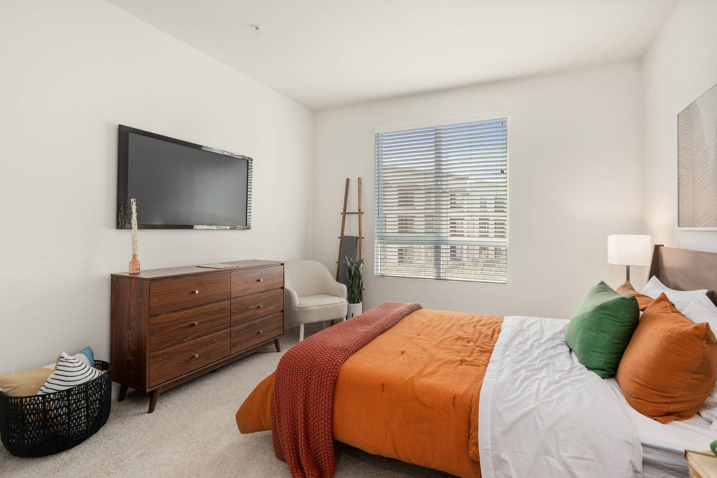 A comfortable bedroom in Alta Upland features a queen-sized bed with orange and green accents, a flat-screen TV, and a sunny window with views of the neighborhood.