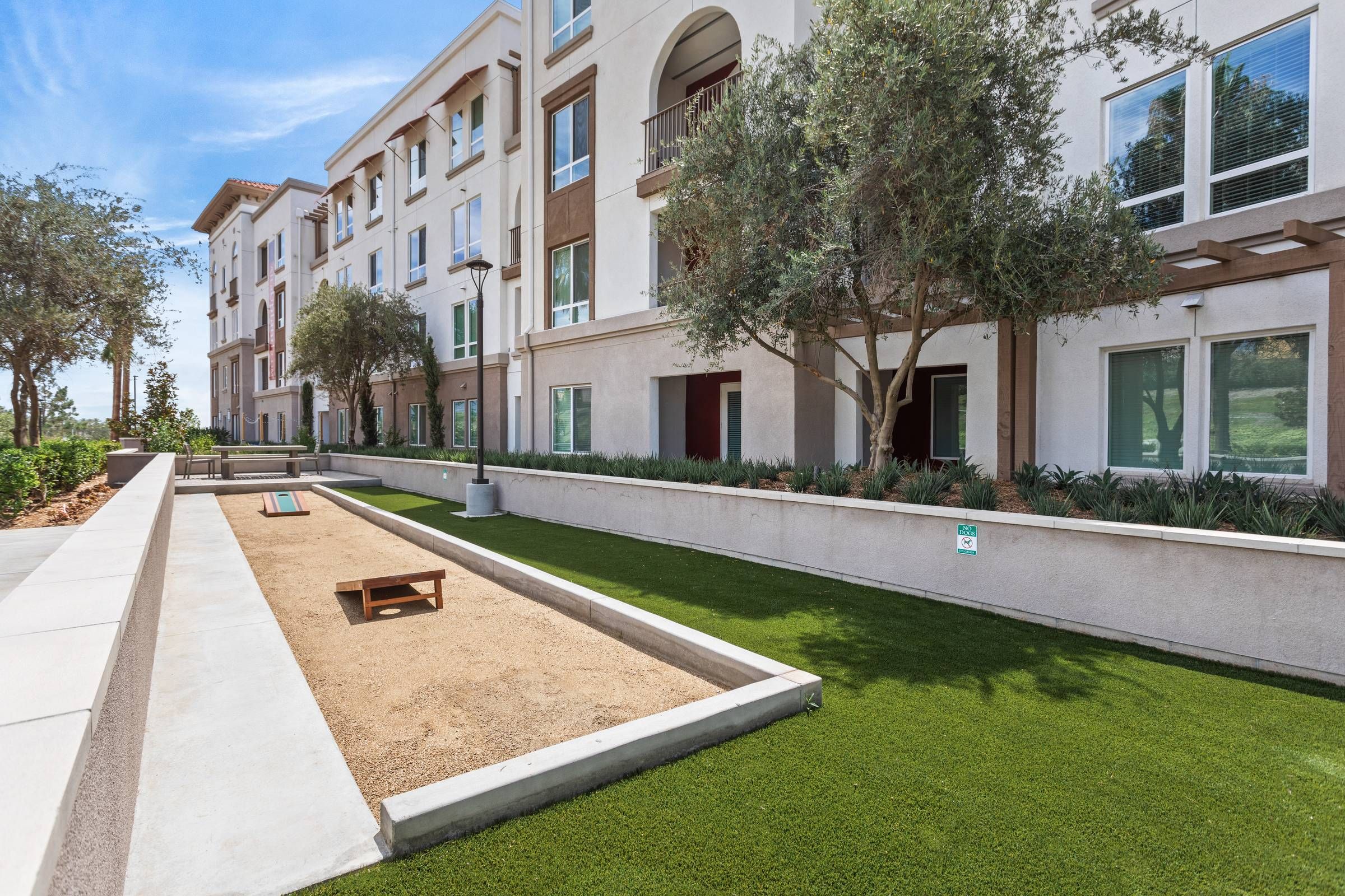 A serene bocce ball court at Alta Upland lies adjacent to a landscaped walkway with neatly trimmed hedges and apartment windows overlooking the area.