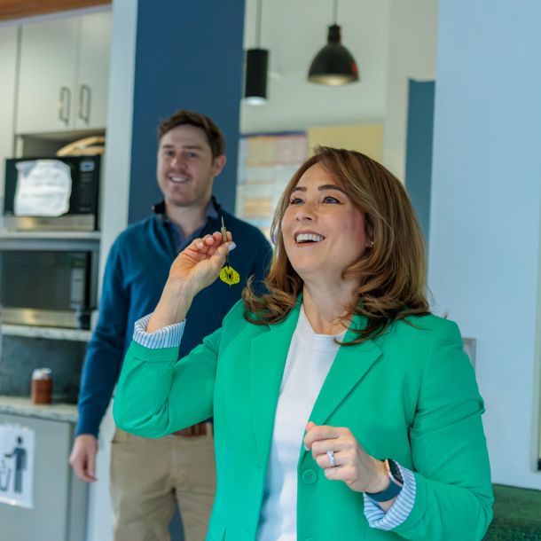 Three people stand in a kitchen area, one woman in a green blazer is smiling and blowing bubbles.